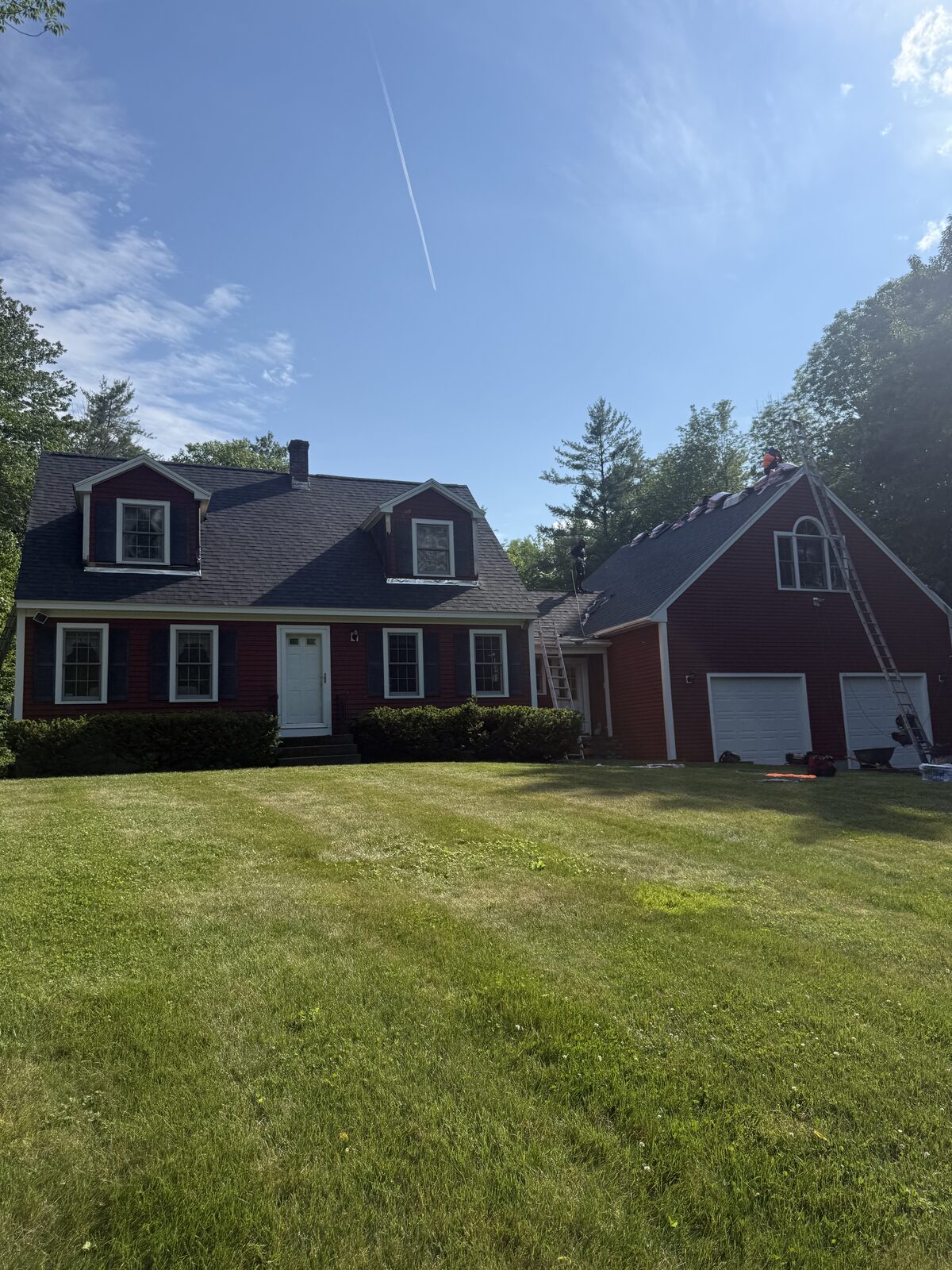Finished dark grey shingle roof on a red cape with three dormers, while the attached garage roof is still being shingled