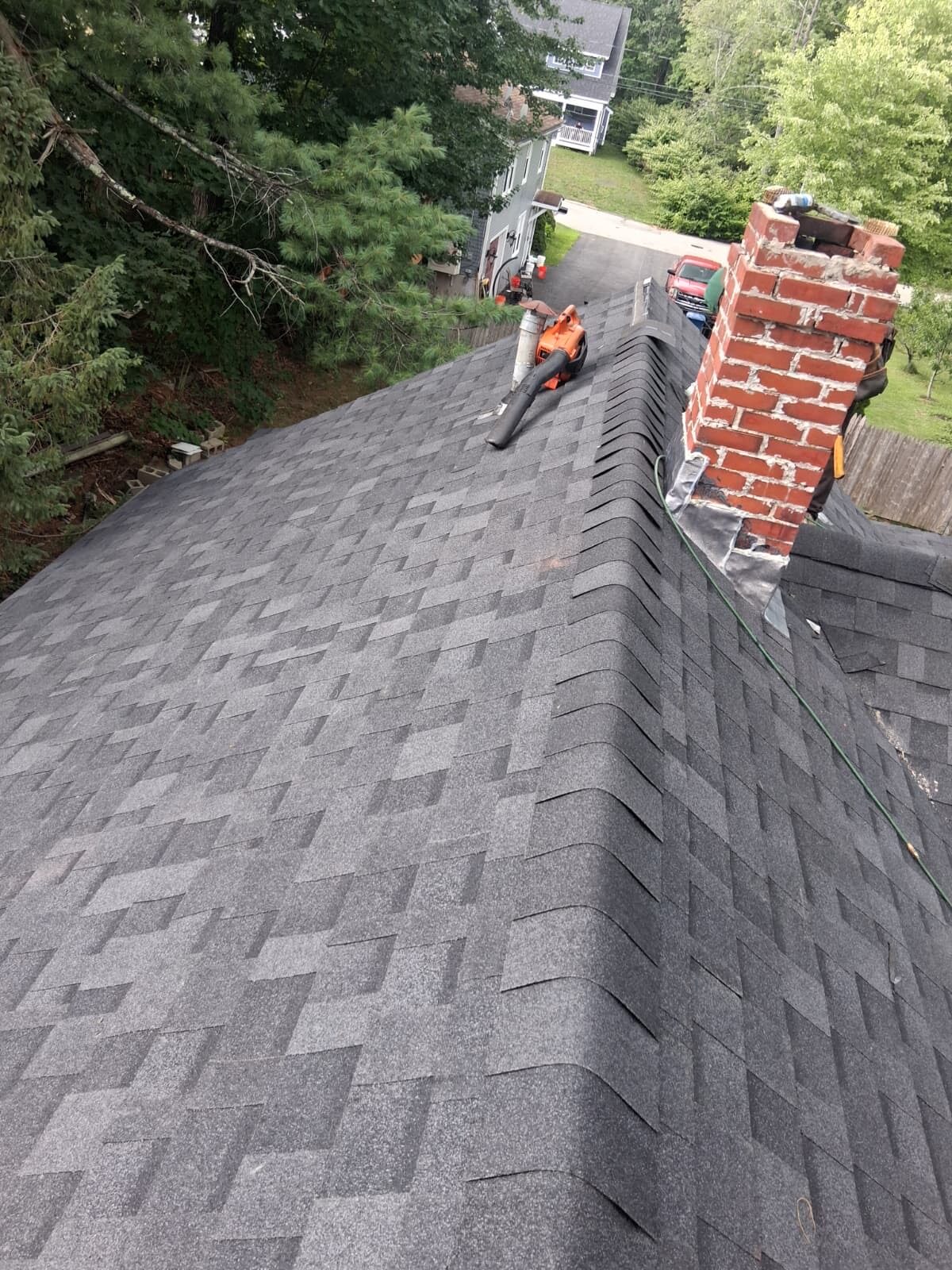 Finished grey architectural shingle roof seen along the ridge, with new ridge cap shingles and fresh metal flashing worked around a brick chimney