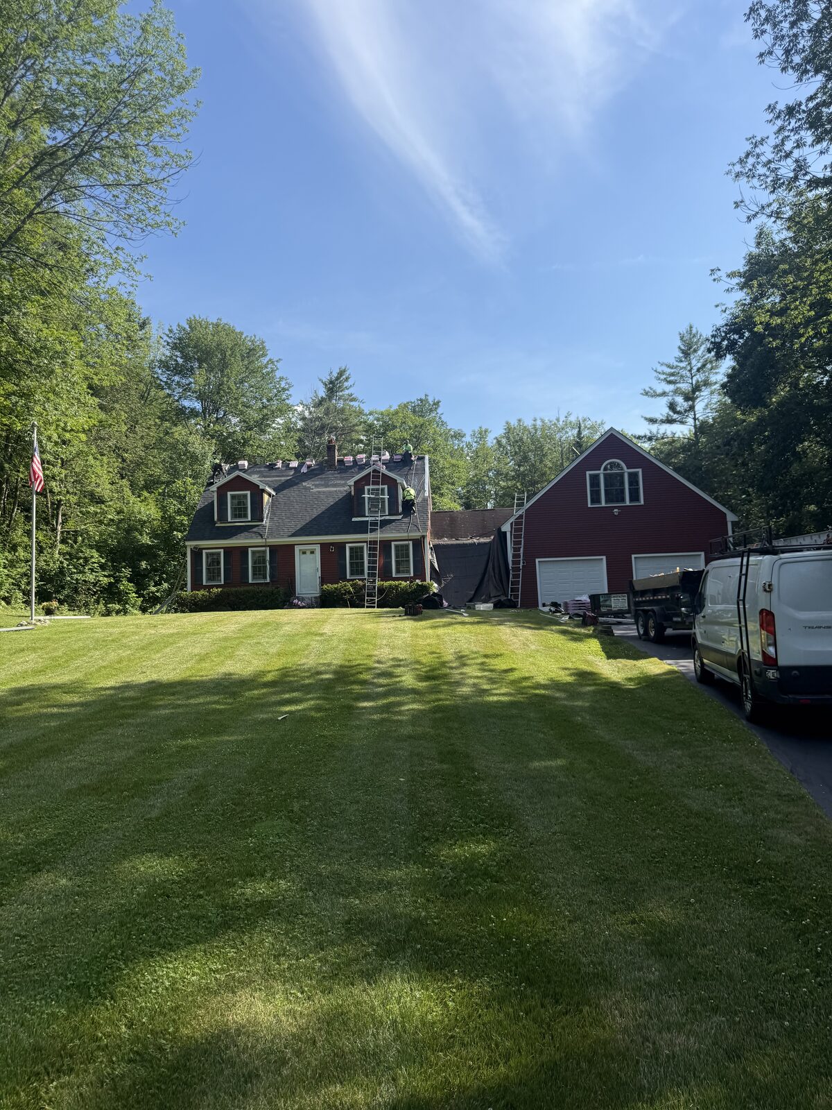 Shingle roof replacement underway on a dormered cape, with ladders at the eaves, bundles staged along the ridge and a crew member working near the chimney