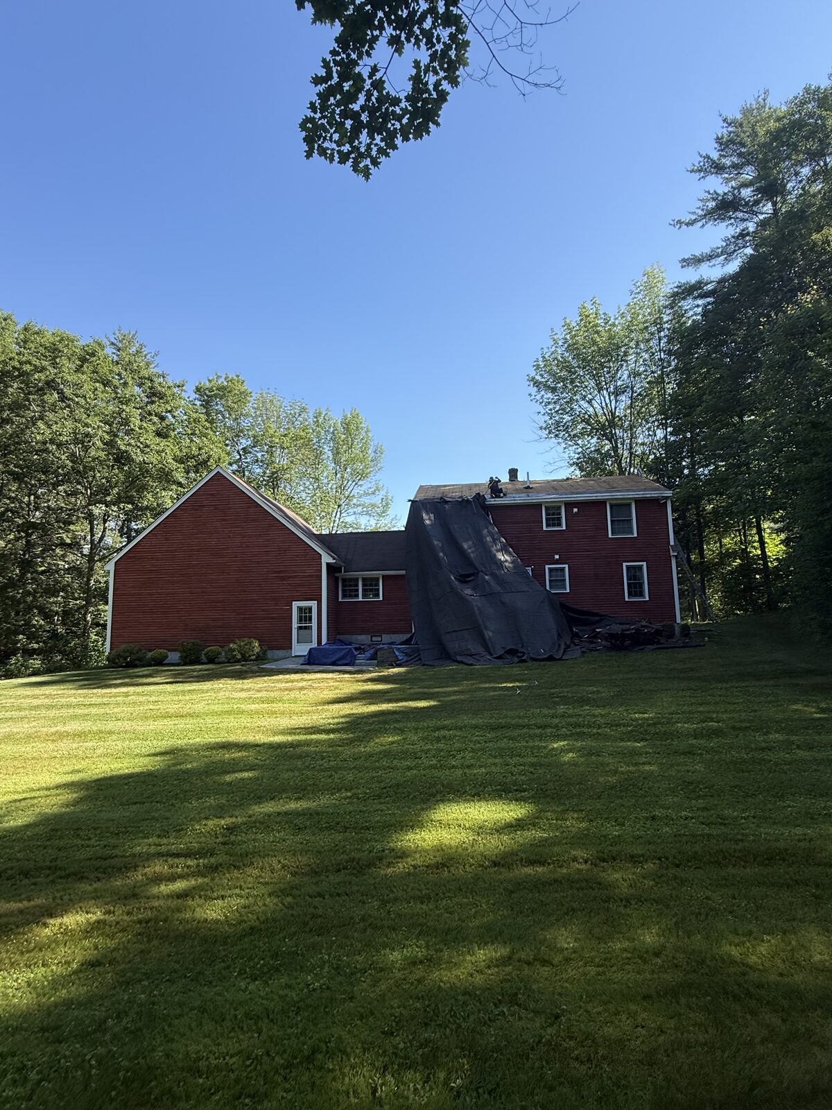 Large tarp hung from the rear roof slope of a red clapboard house to catch stripped shingles and protect the siding and plantings below