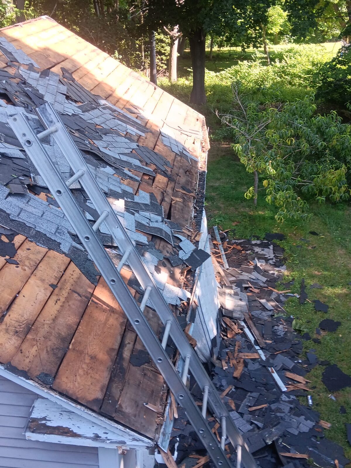 Close view of worn shingles peeled back to bare wood plank decking along a roof edge, with an extension ladder set on the slope and debris on the ground below